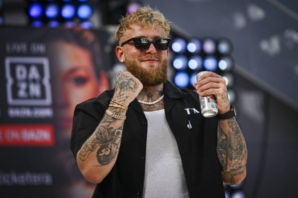 Jake Paul in a black shirt and white vest holds his fists aloft while holding a Celsius can and wearing sunglasses