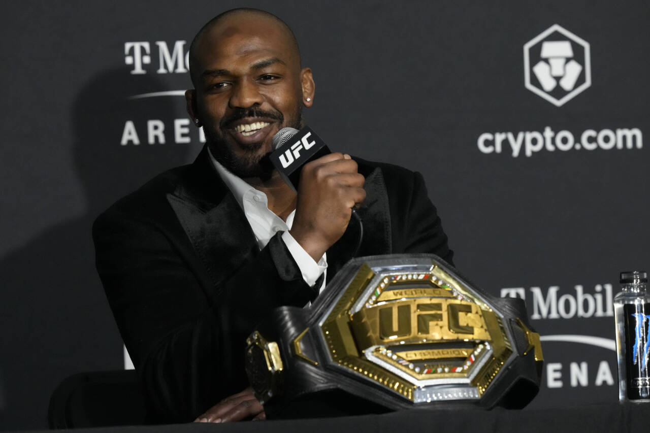 Jon Jones, smiling in a black suit, holds a UFC-branded microphone while his UFC title belt sits on a table in front of him