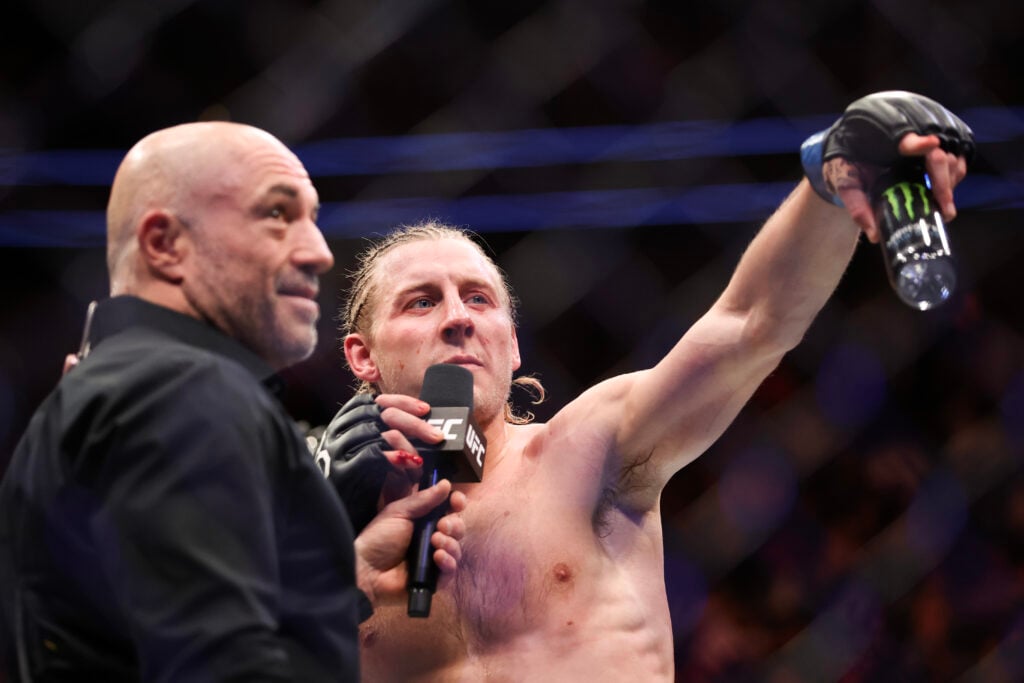 Joe Rogan wearing all black holds a microphone with 'UFC' on it for Paddy Pimblett who is shirtless and speaking while holding a Monster Energy