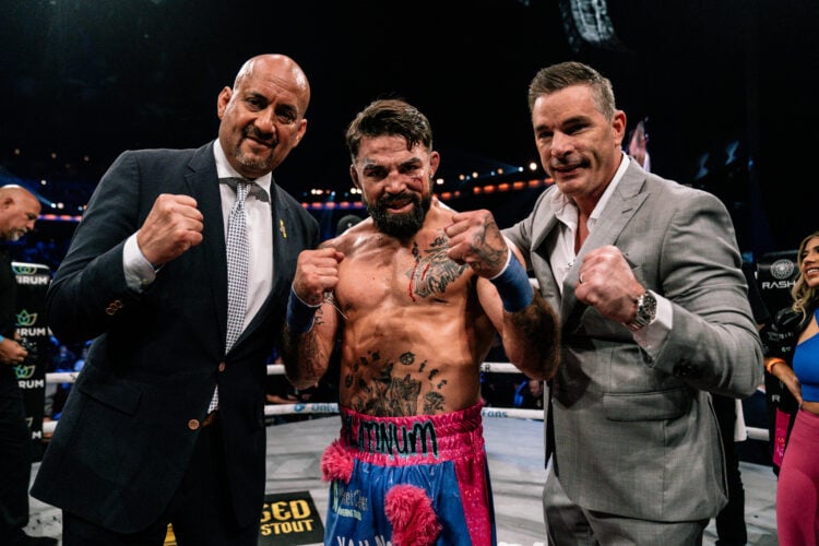 Mike Perry shirtless in pink and blue shorts that read 'Platinum' stands between two men in suits