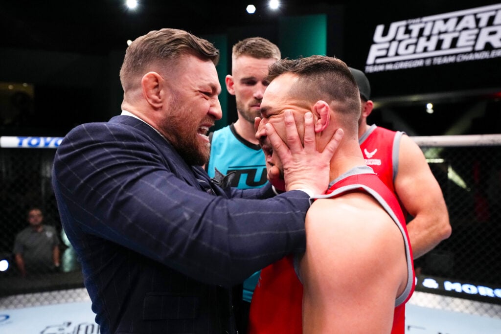 Conor McGregor in a suit pushes Michael Chandler in a red sleeveless shirt