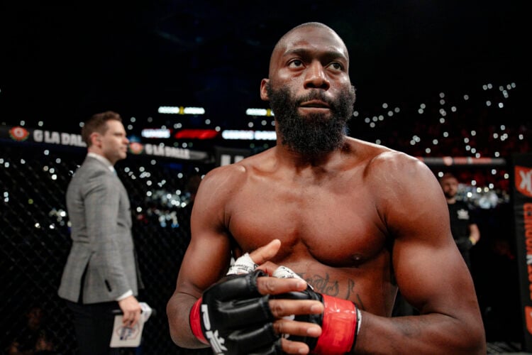 Cedric Doumbe shirtless wearing PFL MMA gloves with red wraps stands in an MMA cage while a crowd light up the venue behind him