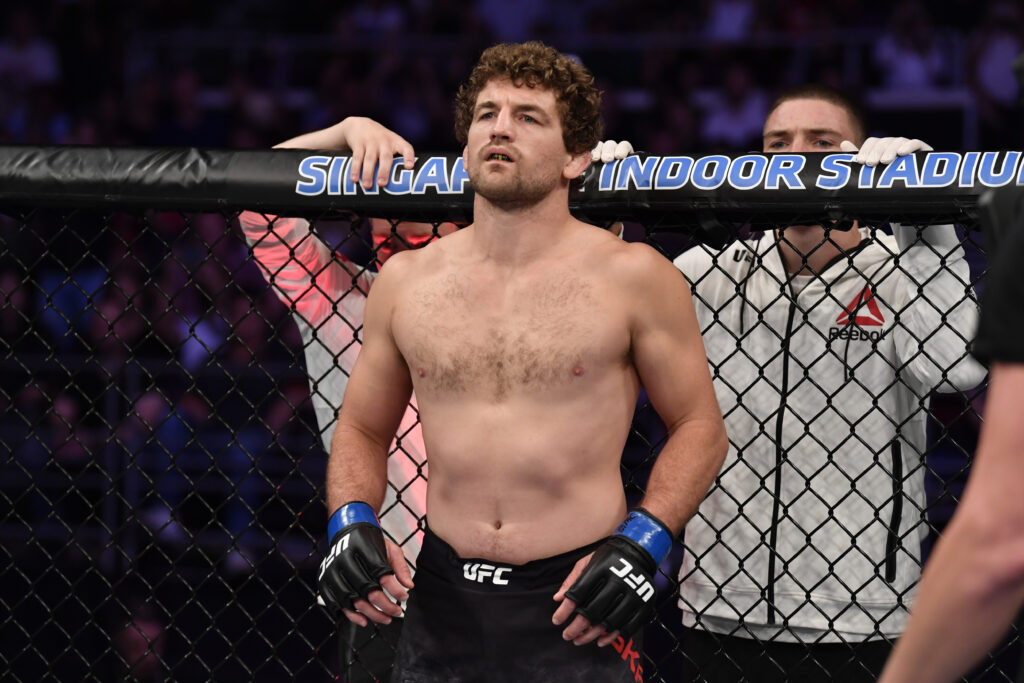Ben Askren shirtless in all UFC gear inside a cage with Singapore Indoor Stadium written on top and a coach behind
