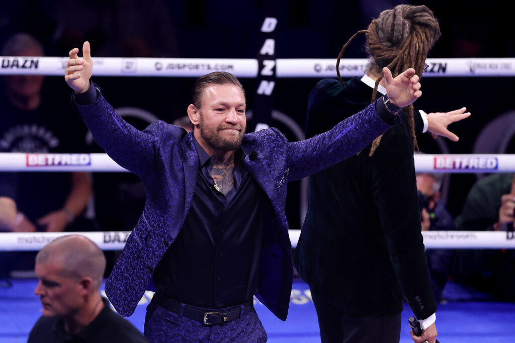 Conor McGregor wearing a blue suit soaks up crowd reaction in a boxing ring