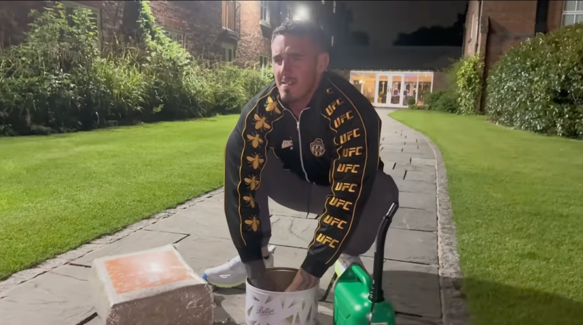 Tom Aspinall with his hands in a bowl purportedly of petrol, with a bail of hay and a bottle of petrol