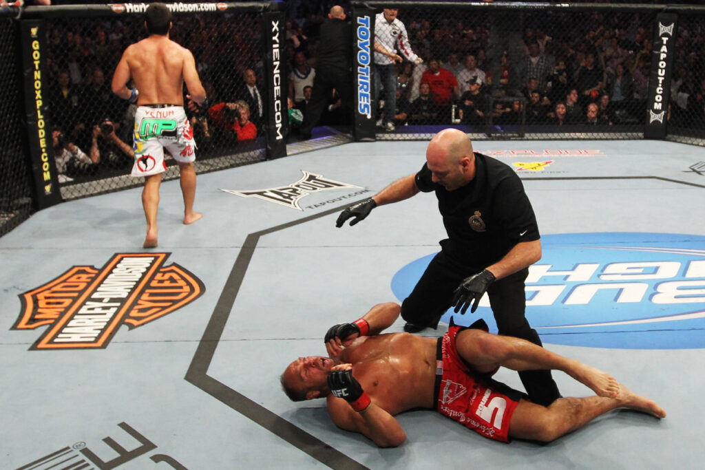 Randy Couture on floor as Lyoto Machida walks off after his KO win at UFC 129