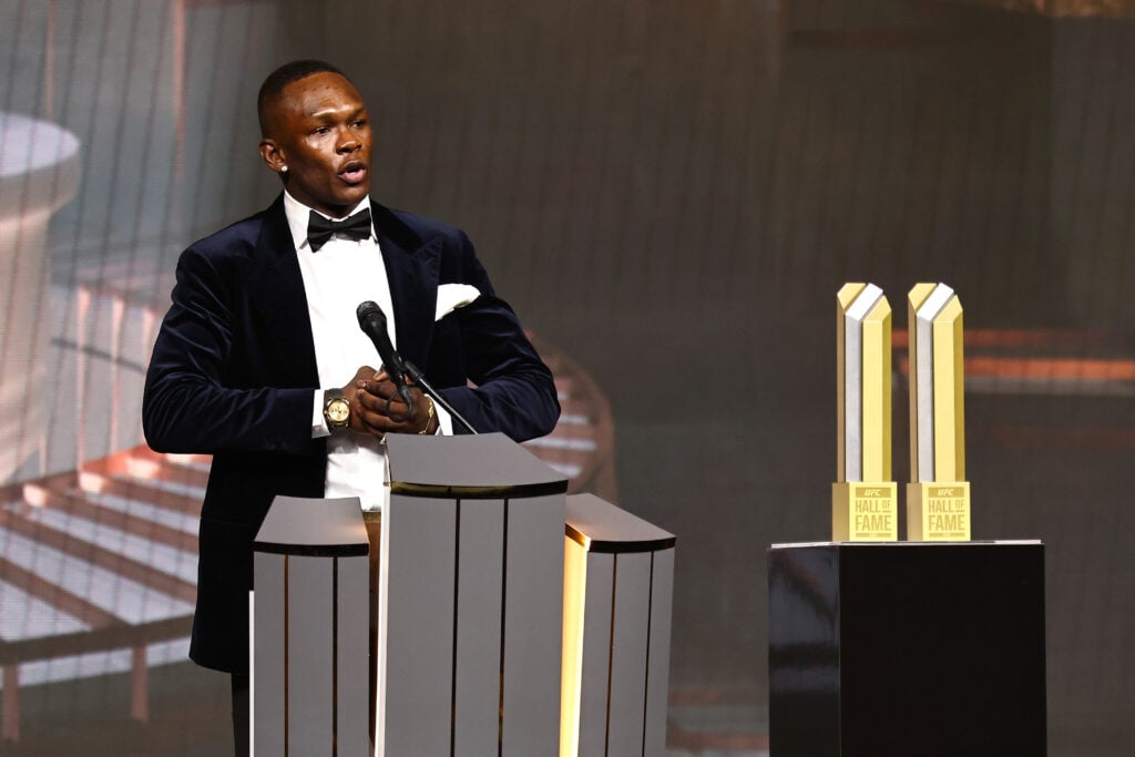 Israel Adesanya accepts his UFC Hall of Fame honor, wearing a black tuxedo on stage next to two Hall of Fame statues