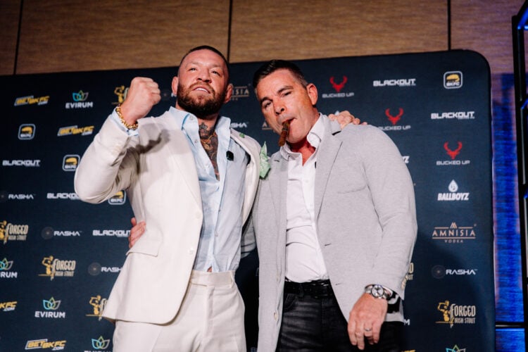Conor McGregor in a white suit poses, fist up, with David Feldman who is smoking a cigar at a BKFC press conference