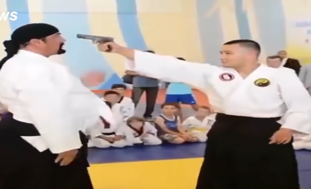Steven Seagal doing an Aikido demonstration with a training partner pointing a gun to his head.