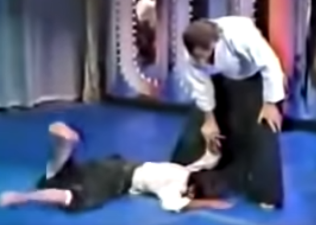 Steven Seagal on the Merv Griffin show in 1982, applying strong wristlock on prone opponent who is tapping out.