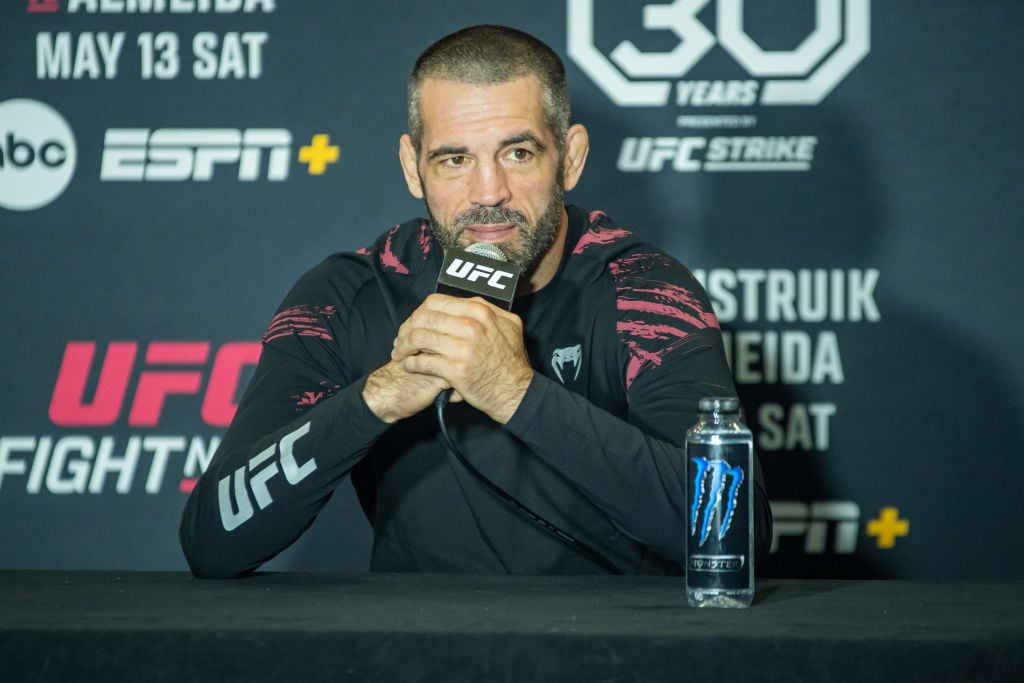 Matt Brown in a black and red tracksuit holds a UFC microphone while leaning on a table. There are advertising boards behind him and there is a Monster-branded water bottle next to him on the table